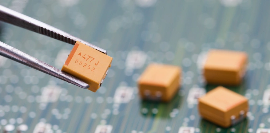Tweezers placing a yellow SMD tantalum capacitor on a PCB, highlighting the need to observe correct polarity during assembly