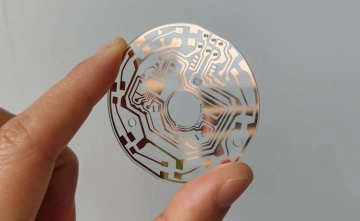 Close-up of a circular transparent PCB with intricate copper circuit traces held between two fingers